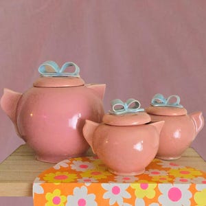 May include: A pink ceramic teapot and two matching lidded sugar bowls, each featuring a light blue bow-shaped handle on the lid. The set has a smooth, glossy finish and rounded bodies, presented together as a cohesive collection of kitchenware.