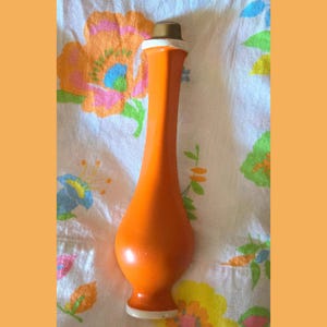 May include: A tall, slender orange vase with a smooth, matte finish. The vase features a bulbous base that tapers into a long, narrow neck, topped with a small, cylindrical metallic cap. The design is simple, vertical, and symmetrical.