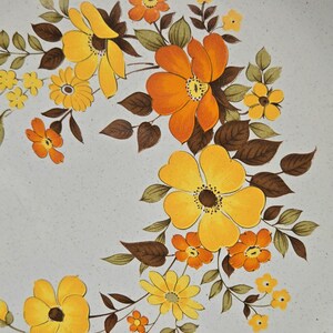 May include: Close-up of a ceramic plate with a floral design. The flowers are in shades of yellow and orange, with brown leaves and stems. The background is a speckled, light gray color.