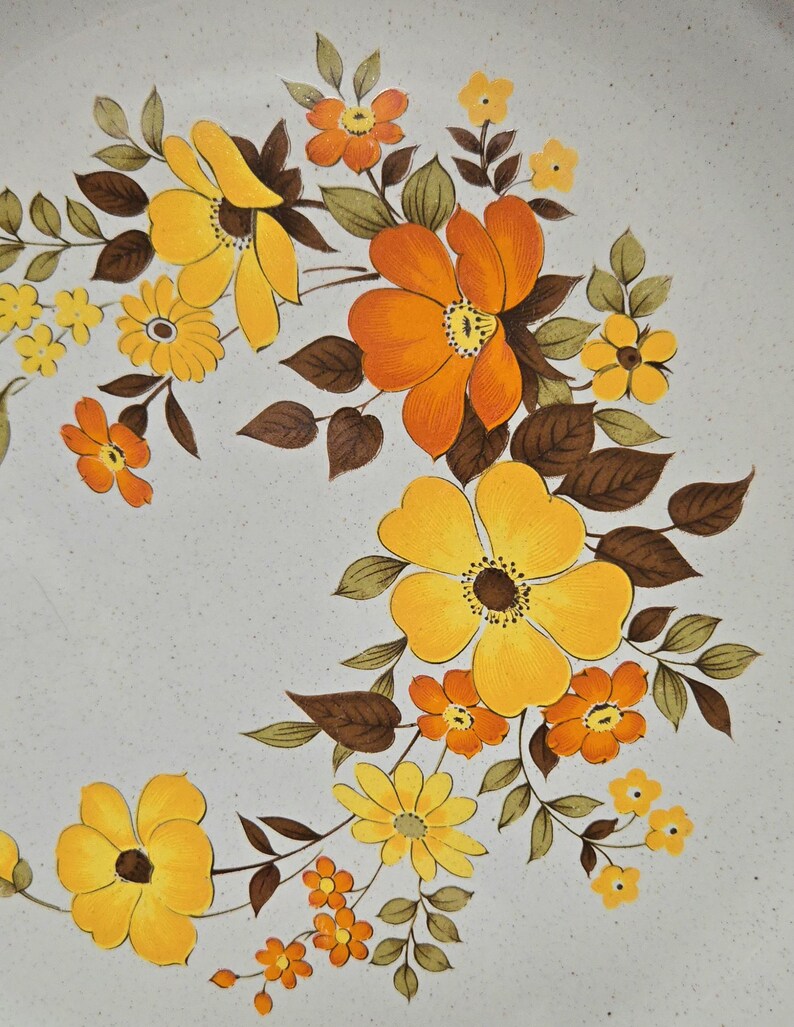 May include: Close-up of a ceramic plate with a floral design. The flowers are in shades of yellow and orange, with brown leaves and stems. The background is a speckled, light gray color.