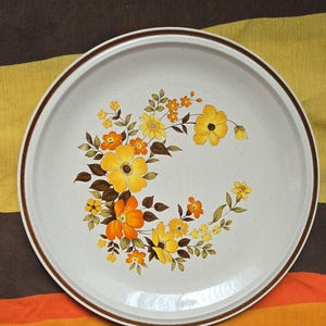 May include: Round, off-white ceramic serving platter with a brown rim and floral design. The floral pattern features yellow and orange flowers with brown leaves. The platter is likely vintage and suitable for serving food.