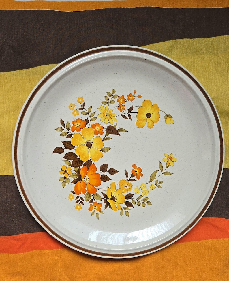 May include: Round, off-white ceramic serving platter with a brown rim and floral design. The floral pattern features yellow and orange flowers with brown leaves. The platter is likely vintage and suitable for serving food.