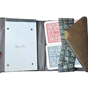 May include: A brown leather folding card game case containing a white score pad with card suit symbols and the text Score Pad, alongside two decks of playing cards held by a black fabric strap featuring a gold geometric pattern and a metal clip.