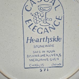 May include: A white stoneware plate with a blue rim and speckled surface. The plate features a blue graphic with the words "CASUAL ELEGANCE" and "Hearthside STONEWARE". The plate is safe for dishwasher, oven, and microwave use.