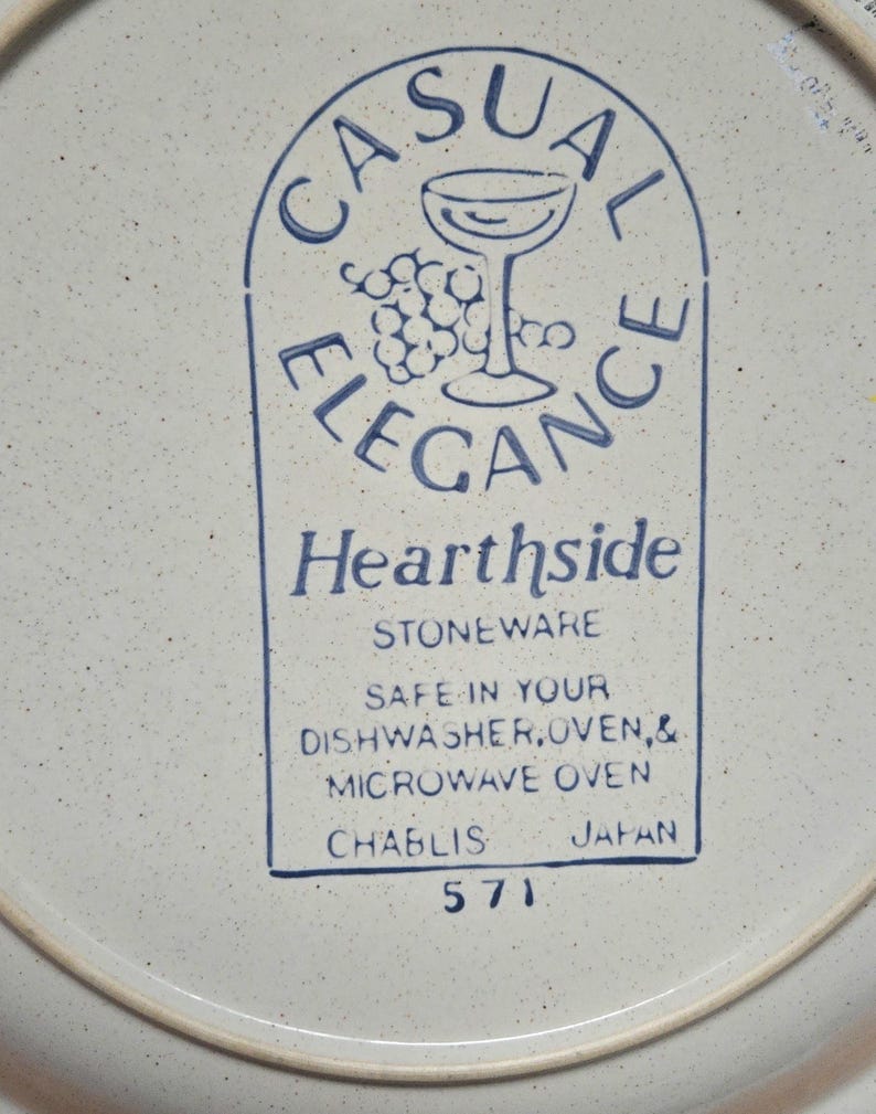 May include: A white stoneware plate with a blue rim and speckled surface. The plate features a blue graphic with the words "CASUAL ELEGANCE" and "Hearthside STONEWARE". The plate is safe for dishwasher, oven, and microwave use.
