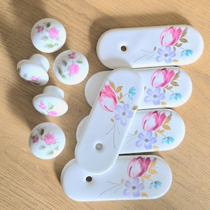 May include: A collection of white ceramic cabinet knobs and pulls. The knobs are round and the pulls are oval, all decorated with pink roses and other floral designs. The hardware is set against a light brown wooden surface.