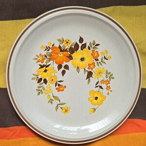May include: A round, off-white ceramic plate with a brown rim and a floral design. The flowers are yellow and orange with brown leaves. The plate has a speckled texture.