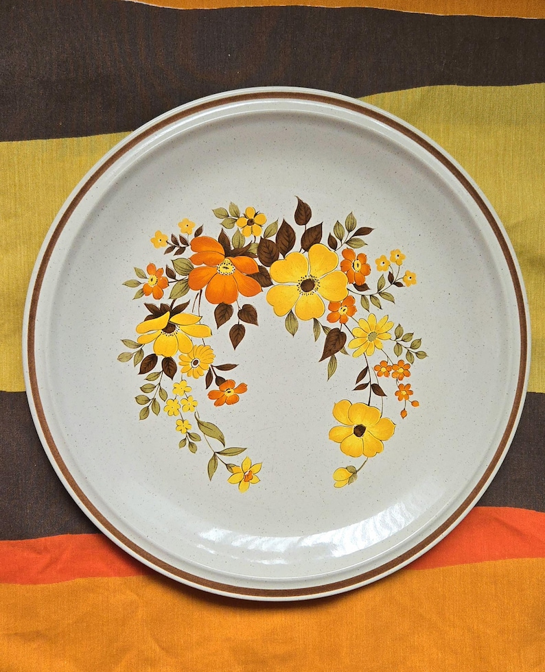 May include: A round, off-white ceramic plate with a brown rim and a floral design. The flowers are yellow and orange with brown leaves. The plate has a speckled texture.