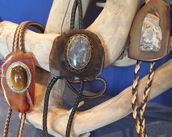 May include: Three bolo ties with unique stone pendants and braided leather cords. The pendants feature a tiger's eye, a gray stone, and a patterned stone. The cords are black, brown, and a mix of tan and brown. The bolo tips are silver and gold.