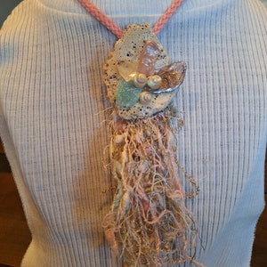May include: A handmade necklace featuring a pendant with a coral-like base, adorned with seashells, crystals, and a cluster of colorful threads. The necklace has a pink braided cord.