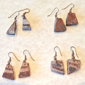 May include: Four pairs of handmade earrings. Each earring features a unique, irregularly shaped stone pendant in shades of gray, brown, and white. The earrings have gold-colored ear wires and are displayed on a light-colored surface.