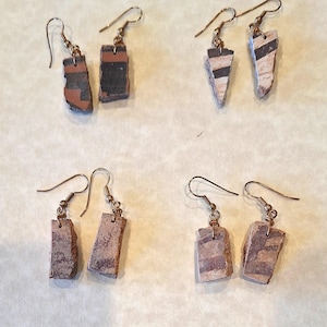 May include: Four pairs of handcrafted earrings. Each pair features a unique, rectangular or triangular stone pendant with brown, black, and white striped patterns. The earrings have gold-colored hooks.