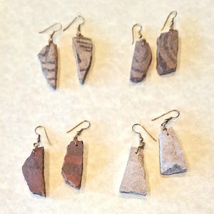 May include: Handmade earrings featuring unique, irregularly shaped stone pendants in shades of brown, gray, and red. Each pair has a different pattern and texture, suspended from gold-colored ear wires. The earrings are displayed on a neutral background.