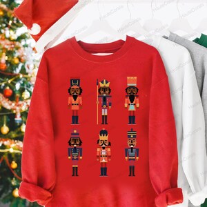 May include: Red Christmas sweatshirt with a design of six black nutcracker figures in various poses. The nutcrackers are wearing colorful hats and uniforms.