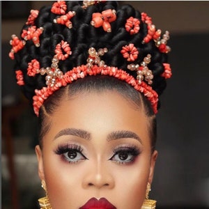 May include: A woman with dark hair styled in a bun adorned with coral beads and gold accents. She is wearing coral bead jewelry around her neck and earrings. She has a red lip and dramatic eye makeup.