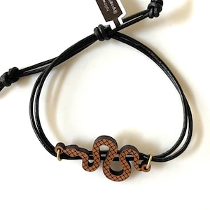 Könnte beinhalten: Ein schwarzes Kordelarmband mit einem Schlangenanhänger aus Holz. Die Schlange ist braun mit einem gemusterten Design. Das Armband hat verstellbare Knoten und kleine goldfarbene Metallringe. Ein kleines Etikett mit Text ist angebracht.