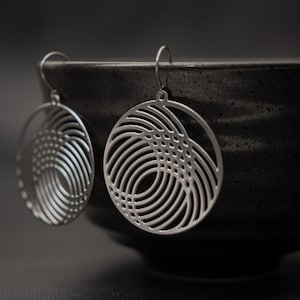 Puede incluir: Pendientes calados plateados con diseño circular. Los pendientes presentan círculos concéntricos y un patrón tejido, suspendidos de un simple gancho. Los pendientes se exhiben sobre un fondo oscuro.