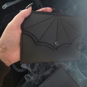 May include: Black leather wallet with a bat wing design on the flap, secured by a snap closure. The wallet is held in a person's hands, showcasing its sleek, gothic aesthetic. A second, smaller black wallet is also visible.