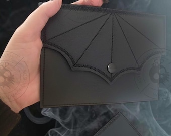 May include: A black leather wallet featuring a bat wing design on the flap, fastened with a press stud. The wallet is held in a person's hands, highlighting its gothic style. A second, smaller black wallet is also visible.
