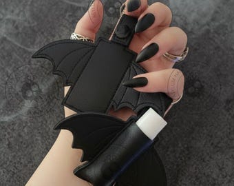 May include: Black leather bat-shaped keychain and lip balm holder. The keychain features bat wings and a small loop for keys. The lip balm holder is also bat-shaped, with a white lip balm tube inside. The background is a dark gray.