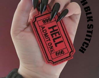 May include: A red ticket with black stitching that reads "HELL ADMIT ONE 666" is held in a hand with long, black, pointed fingernails. The text "RED WITH BLK STITCH" is arched above the ticket.