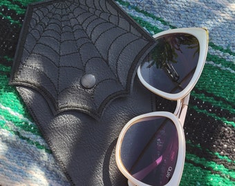 May include: Black leather sunglasses case with a stitched spiderweb design and a snap closure. White sunglasses with a gold frame and dark lenses rest on the case. The sunglasses have the word "Quay" printed on the lens.