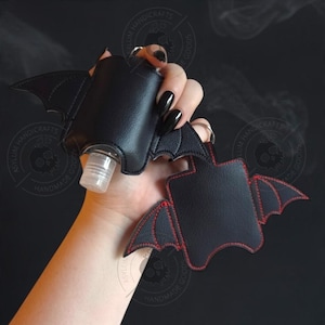 Puede incluir: Dos fundas de piel sint&eacute;tica negra con forma de murci&eacute;lago y alas puntiagudas. Una funda contiene una peque&ntilde;a botella de pl&aacute;stico transparente con tap&oacute;n blanco. La otra tiene costuras rojas visibles en los bordes y un anillo met&aacute;lico.