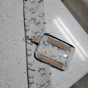 K&ouml;nnte beinhalten: Eine wei&szlig;e Stofftasche mit Rei&szlig;verschluss und einem transparenten Plastikfenster. Die Tasche ist mit einem sich wiederholenden Muster von Eulen verziert, die Briefe tragen. Die Tasche ist an einem Schl&uuml;sselanh&auml;nger mit einem silbernen Ring befestigt.