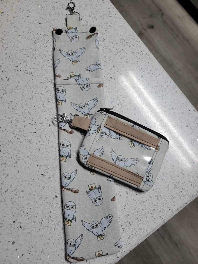 K&ouml;nnte beinhalten: Eine wei&szlig;e Stofftasche mit Rei&szlig;verschluss und einem transparenten Plastikfenster. Die Tasche ist mit einem sich wiederholenden Muster von Eulen verziert, die Briefe tragen. Die Tasche ist an einem Schl&uuml;sselanh&auml;nger mit einem silbernen Ring befestigt.
