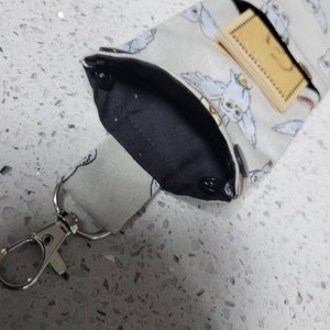K&ouml;nnte beinhalten: Ein graues Stoffband mit einer schwarzen Stofftasche und einem silbernen Metallverschluss. Die Tasche hat einen wei&szlig;en Eulen-Druck und eine kleine braune Karte im Inneren.