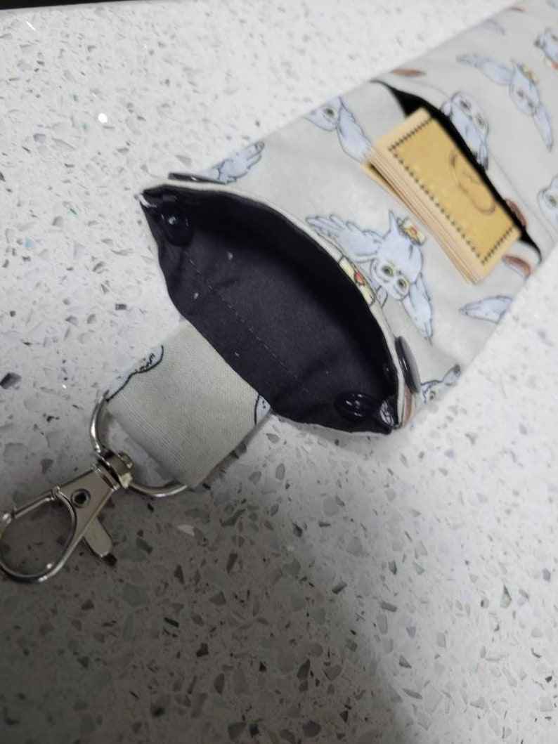 K&ouml;nnte beinhalten: Ein graues Stoffband mit einer schwarzen Stofftasche und einem silbernen Metallverschluss. Die Tasche hat einen wei&szlig;en Eulen-Druck und eine kleine braune Karte im Inneren.