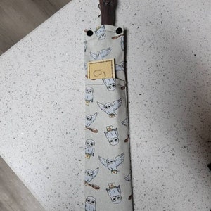 K&ouml;nnte beinhalten: Ein brauner Holzstab mit einem geschnitzten Griff, in einer grauen Stofftasche mit einem wei&szlig;en Eulenmuster. Die Tasche hat ein kleines braunes Etikett mit einem wei&szlig;en Eulenlogo.