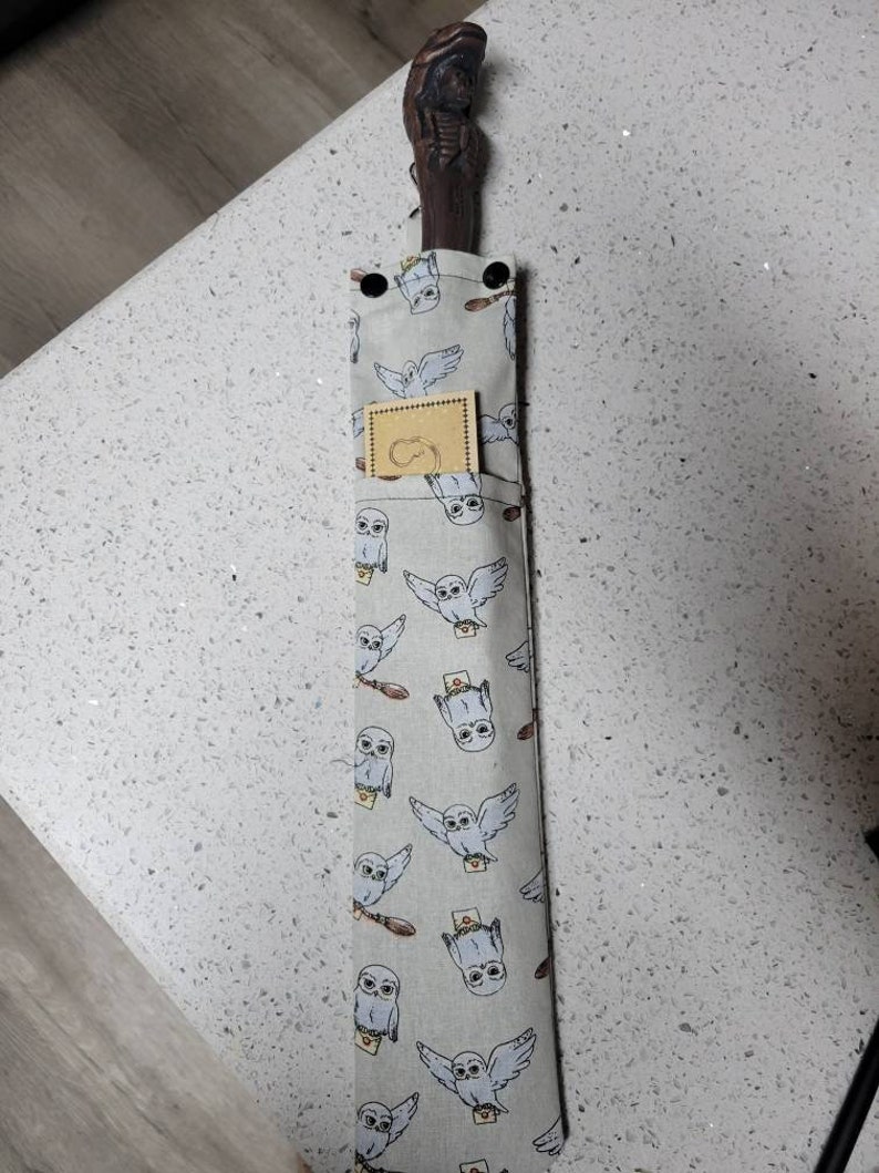 K&ouml;nnte beinhalten: Ein brauner Holzstab mit einem geschnitzten Griff, in einer grauen Stofftasche mit einem wei&szlig;en Eulenmuster. Die Tasche hat ein kleines braunes Etikett mit einem wei&szlig;en Eulenlogo.