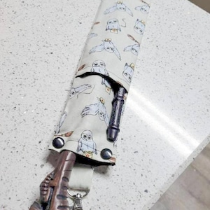 May include: A white fabric pouch with a pattern of owls and broomsticks. The pouch has two black buttons and a brown and black wand is sticking out of the top. A brown wand with a carved handle is attached to a silver carabiner.