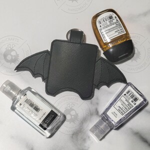 Puede incluir: Un soporte de piel sint&eacute;tica negra para desinfectante de manos con forma de alas de murci&eacute;lago y un anillo de metal en la parte superior. El soporte est&aacute; rodeado por tres frascos de pl&aacute;stico rectangulares con gel transparente y &aacute;mbar.