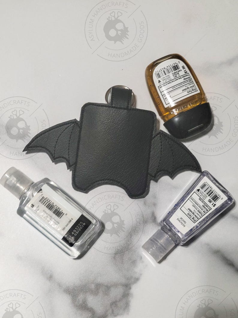 Puede incluir: Un soporte de piel sint&eacute;tica negra para desinfectante de manos con forma de alas de murci&eacute;lago y un anillo de metal en la parte superior. El soporte est&aacute; rodeado por tres frascos de pl&aacute;stico rectangulares con gel transparente y &aacute;mbar.