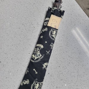 K&ouml;nnte beinhalten: Eine schwarze Stoff-Zauberstabtasche mit einem wei&szlig;en Hogwarts-Wappenmuster. Die Tasche hat einen Rei&szlig;verschluss und eine kleine Tasche auf der Vorderseite. Die Tasche ist mit einem Zauberstab aus Holz gef&uuml;llt, der einen sch&auml;delf&ouml;rmigen Griff hat.