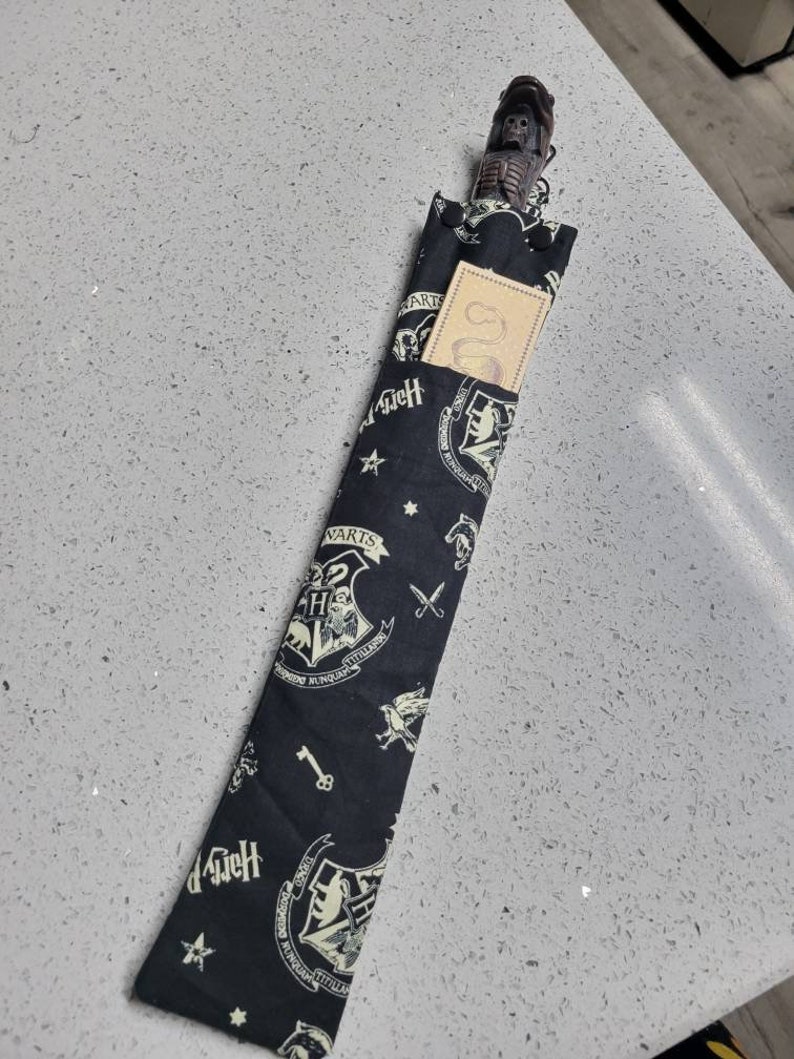 K&ouml;nnte beinhalten: Eine schwarze Stoff-Zauberstabtasche mit einem wei&szlig;en Hogwarts-Wappenmuster. Die Tasche hat einen Rei&szlig;verschluss und eine kleine Tasche auf der Vorderseite. Die Tasche ist mit einem Zauberstab aus Holz gef&uuml;llt, der einen sch&auml;delf&ouml;rmigen Griff hat.