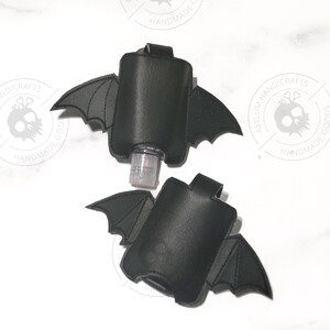 Puede incluir: Dos fundas negras para desinfectante de manos con forma de alas de murci&eacute;lago. Cada funda presenta una bolsa rectangular central con extensiones de alas cosidas a ambos lados y una peque&ntilde;a presilla en la parte superior.