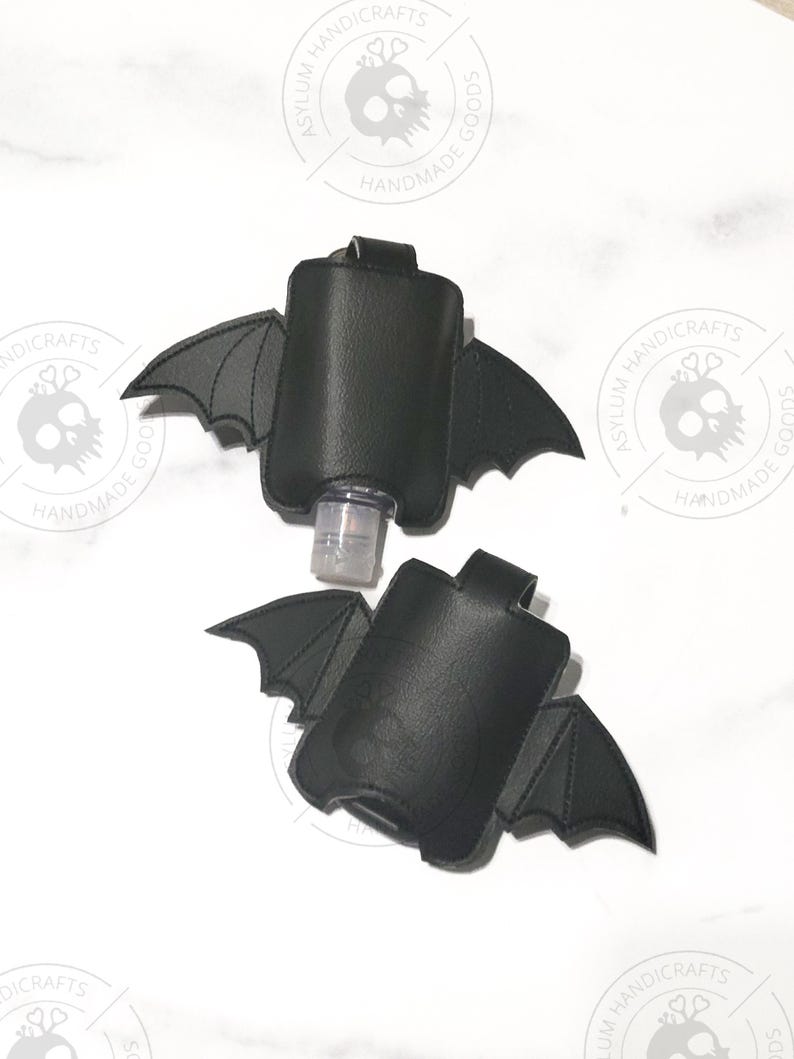 Puede incluir: Dos fundas negras para desinfectante de manos con forma de alas de murci&eacute;lago. Cada funda presenta una bolsa rectangular central con extensiones de alas cosidas a ambos lados y una peque&ntilde;a presilla en la parte superior.