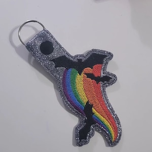 May include: A silver glitter keychain with a rainbow and three black bats. The bats are flying through the rainbow.