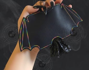 May include: A black bat-shaped dispenser made of textured faux leather with rainbow-colored stitching along the edges. A small black plastic bag is protruding from the bottom center of the bat-shaped holder.