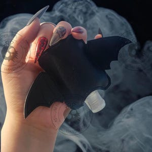 Puede incluir: Un soporte negro con forma de murci&eacute;lago hecho de material liso, con detalles de alas cosidas y un recorte central que revela una peque&ntilde;a botella de pl&aacute;stico transparente con un tap&oacute;n dosificador blanco para sujetar la botella.