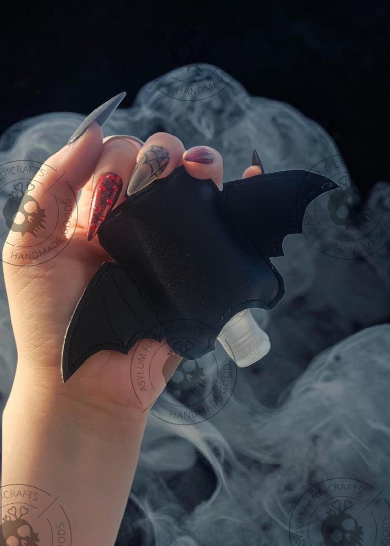 Puede incluir: Un soporte negro con forma de murci&eacute;lago hecho de material liso, con detalles de alas cosidas y un recorte central que revela una peque&ntilde;a botella de pl&aacute;stico transparente con un tap&oacute;n dosificador blanco para sujetar la botella.