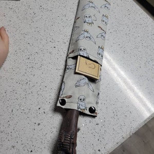 K&ouml;nnte beinhalten: Ein brauner Holzstab mit einem geschnitzten Griff, aufbewahrt in einer grauen Stofftasche mit einem wei&szlig;en Eulenmuster. Die Tasche hat eine kleine Tasche mit einem braunen Papieretikett.