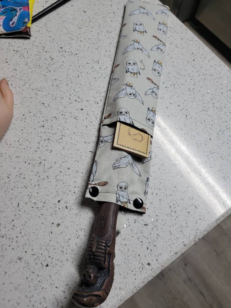 K&ouml;nnte beinhalten: Ein brauner Holzstab mit einem geschnitzten Griff, aufbewahrt in einer grauen Stofftasche mit einem wei&szlig;en Eulenmuster. Die Tasche hat eine kleine Tasche mit einem braunen Papieretikett.