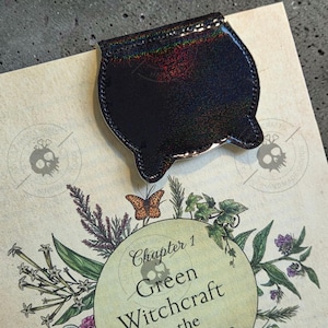 May include: A black holographic cauldron-shaped magnetic bookmark featuring a subtle glitter finish. The bookmark is placed on a page with a botanical illustration of flowers, leaves, and a butterfly, with the text Chapter 1 Green Witchcraft.