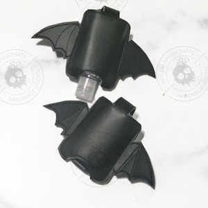 Puede incluir: Dos soportes de piel sint&eacute;tica negra para desinfectante de manos con forma de murci&eacute;lago y alas cosidas. Uno contiene una botella de pl&aacute;stico transparente con dosificador. Cada soporte tiene una trabilla y un logo de calavera.