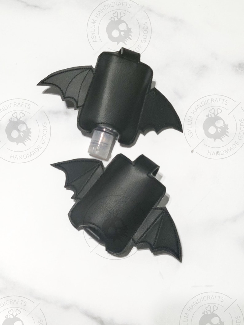 Puede incluir: Dos soportes de piel sint&eacute;tica negra para desinfectante de manos con forma de murci&eacute;lago y alas cosidas. Uno contiene una botella de pl&aacute;stico transparente con dosificador. Cada soporte tiene una trabilla y un logo de calavera.