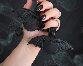 May include: Two black bat-shaped keychains made of a textured material with stitched wing details. Each keychain features a circular top section with a black snap button closure and a metal ring attached for securing keys or other small items.